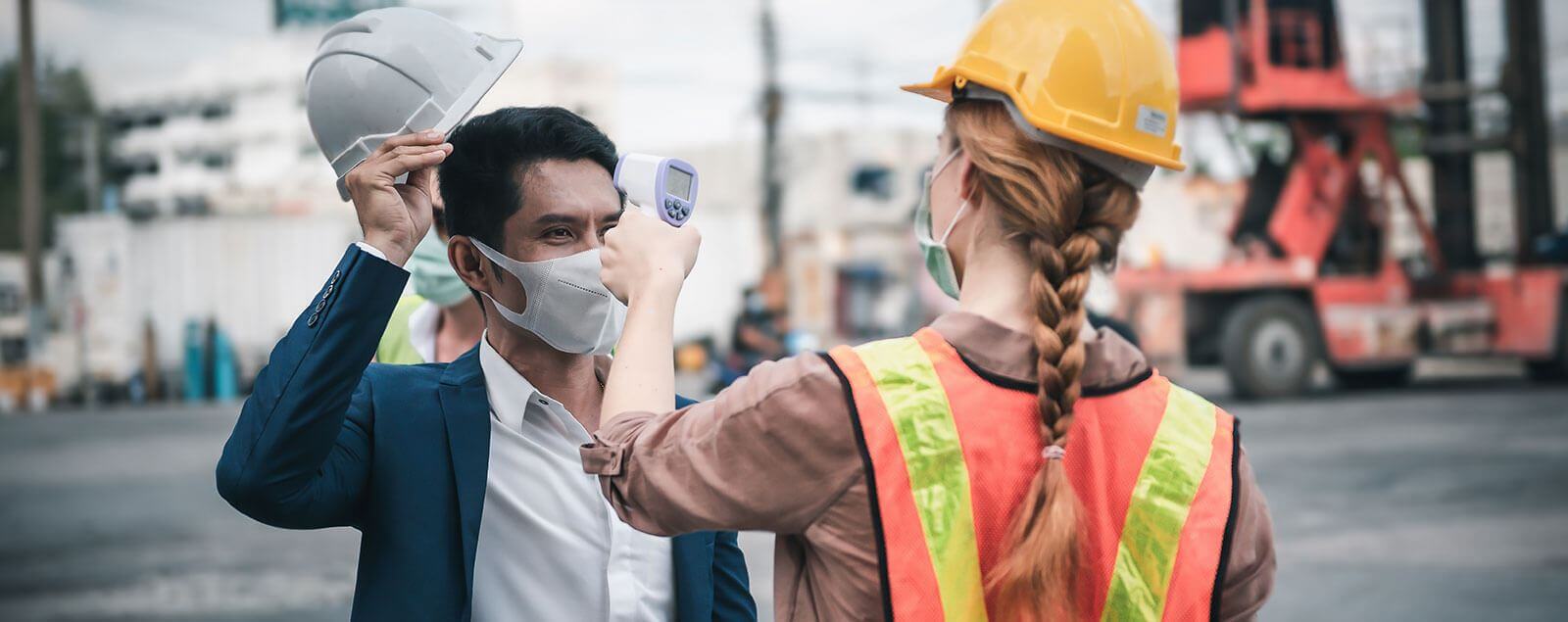 Lady in hardhat and hi-vis taking the temperature of man in suit and hardhat