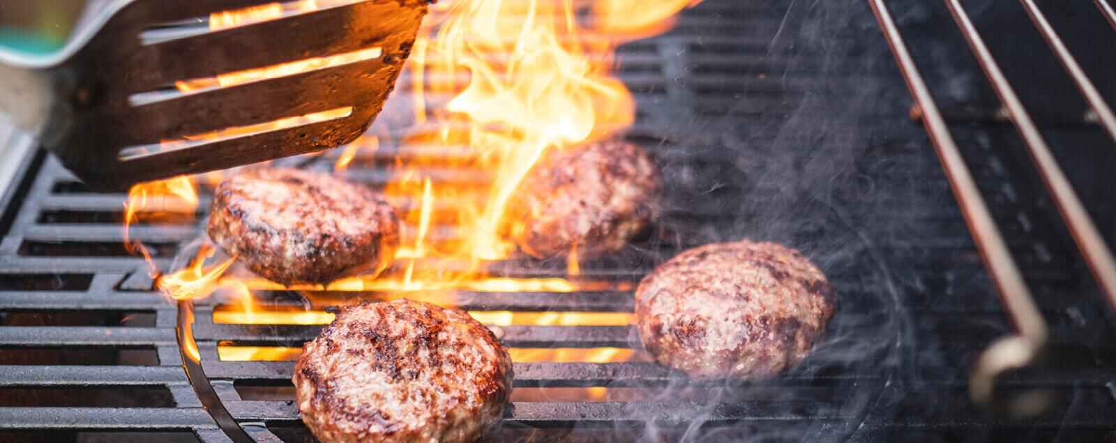 Four burgers cooking on barbeque with flams rising from grill