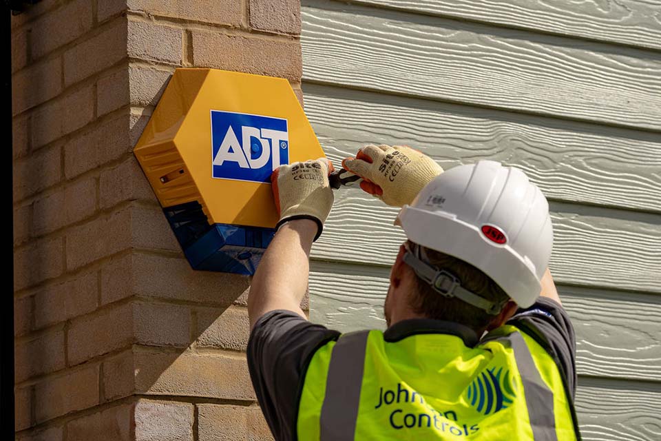 Man in hardhat and hi-vis installing ADT alarm on exterior wall