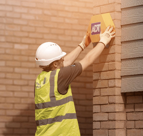 Man in hardhat and hi-vis installing ADT alarm on exterior wall