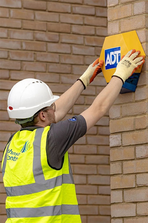 Man in hardhat and hi-vis installing ADT alarm on exterior wall