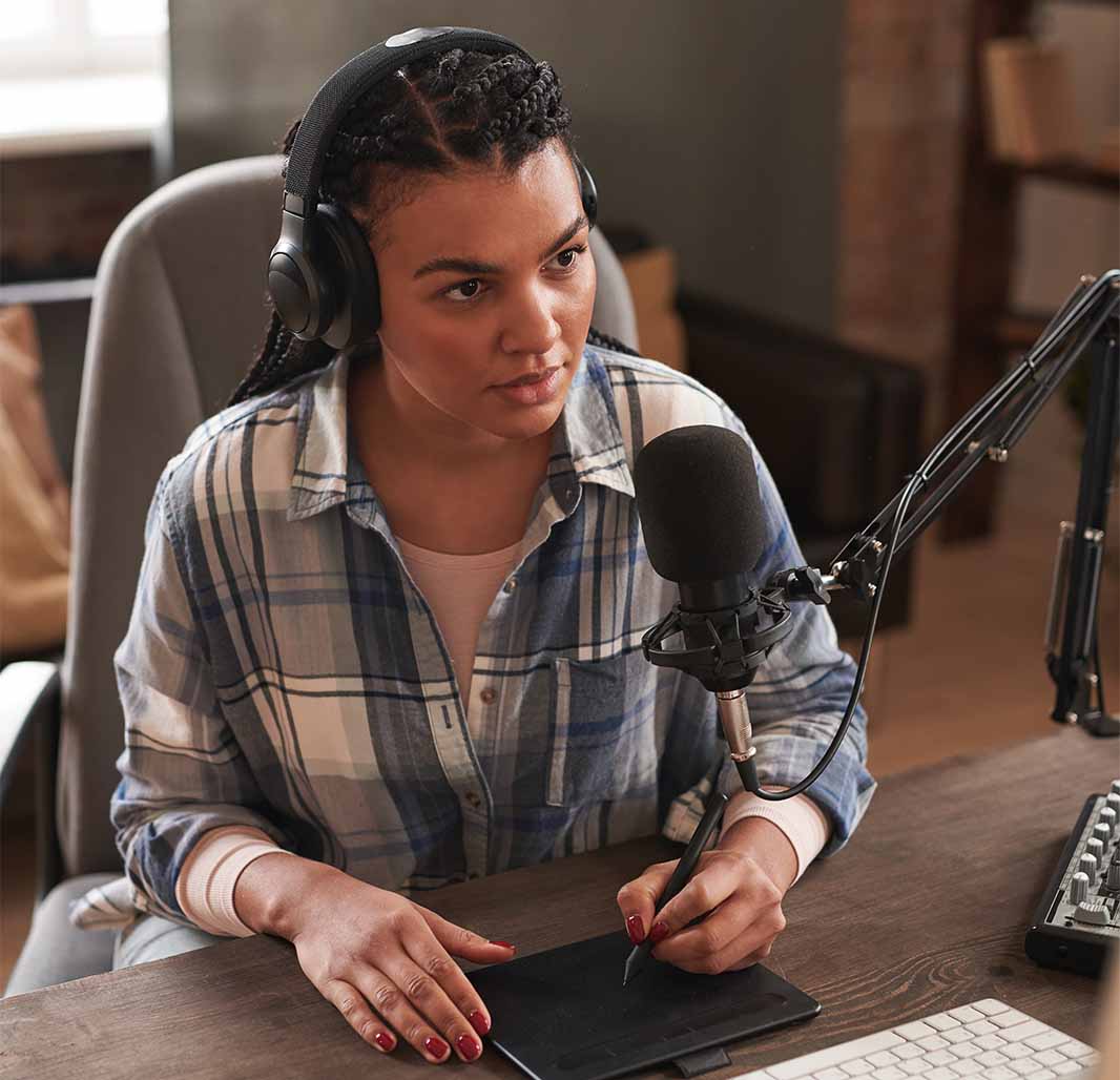 Lady sat at desk positioned next to a microphone, wearing headphones and drawing on a digital tablet