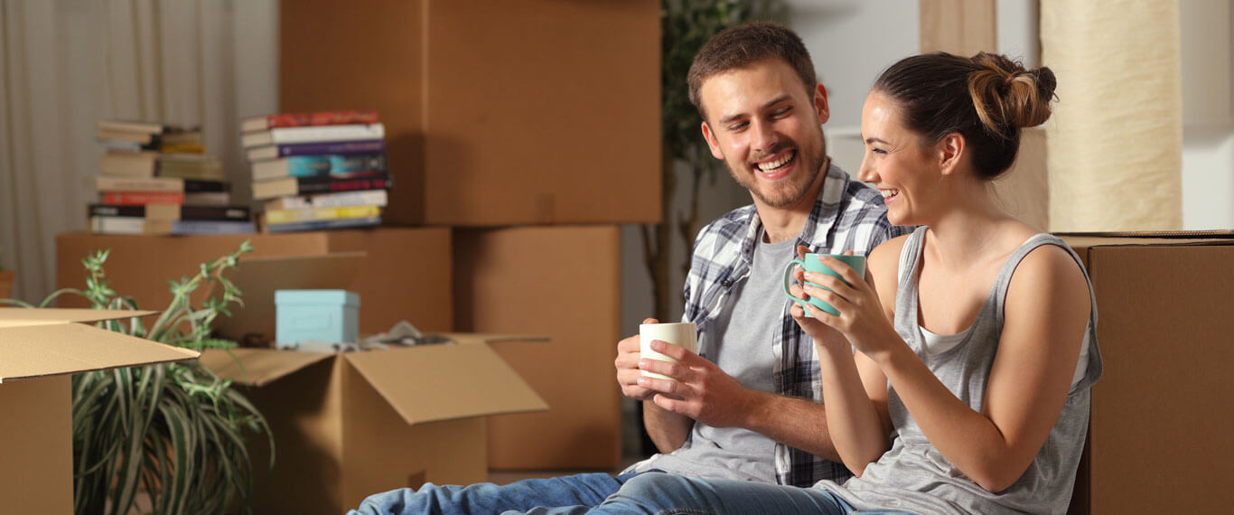 Couple sat down laughing and holding mugs surrounded by boxes